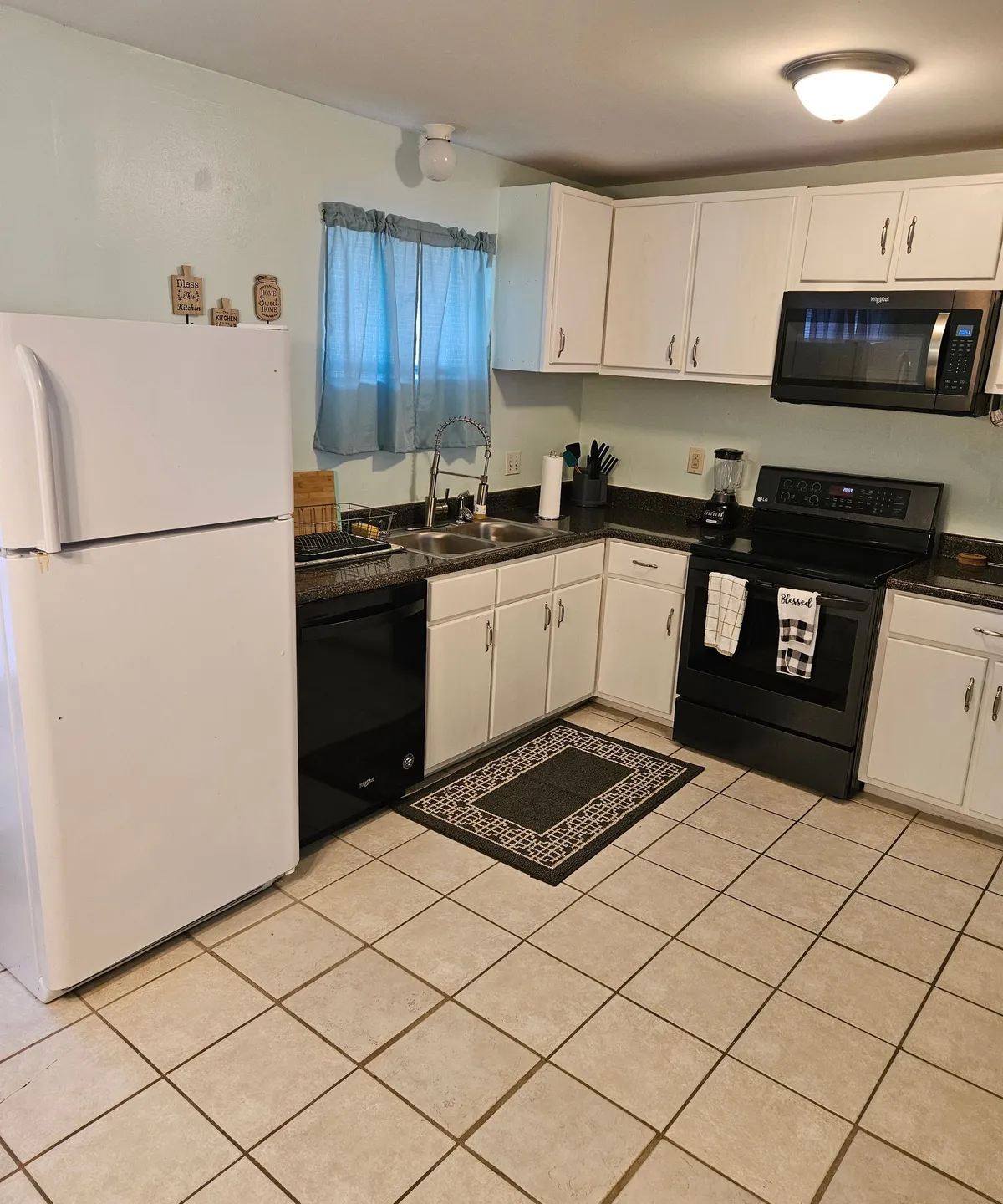 A second angle of the kitchenette highlights both the appliances and the countertop workspace under another curtained window. The layout is efficient, ideal for preparing meals or snacks for a group or family, reinforcing the theme of practical everyday use.