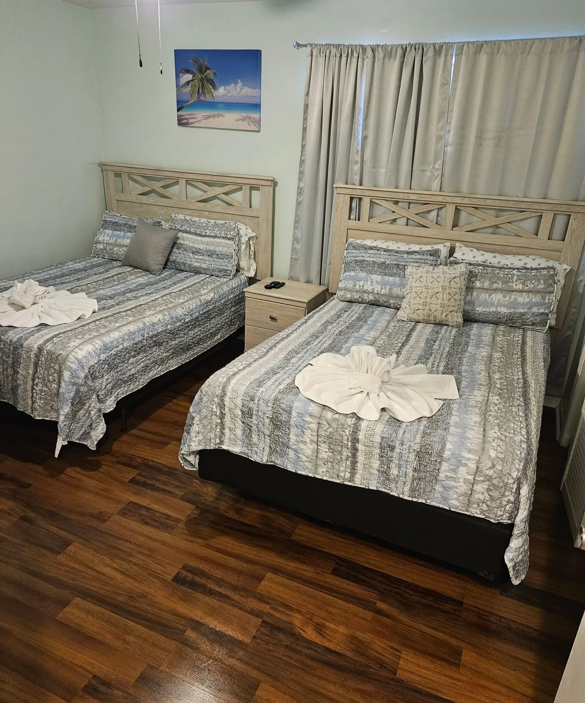 This image displays two neatly made beds set against a backdrop of tropical artwork, with a wooden nightstand between them. The scene is calm and inviting, and the beds suggest room for multiple guests, ideal for families or groups traveling together. Soft lighting and colorful decor evoke a sense of relaxation after a day of exploration.