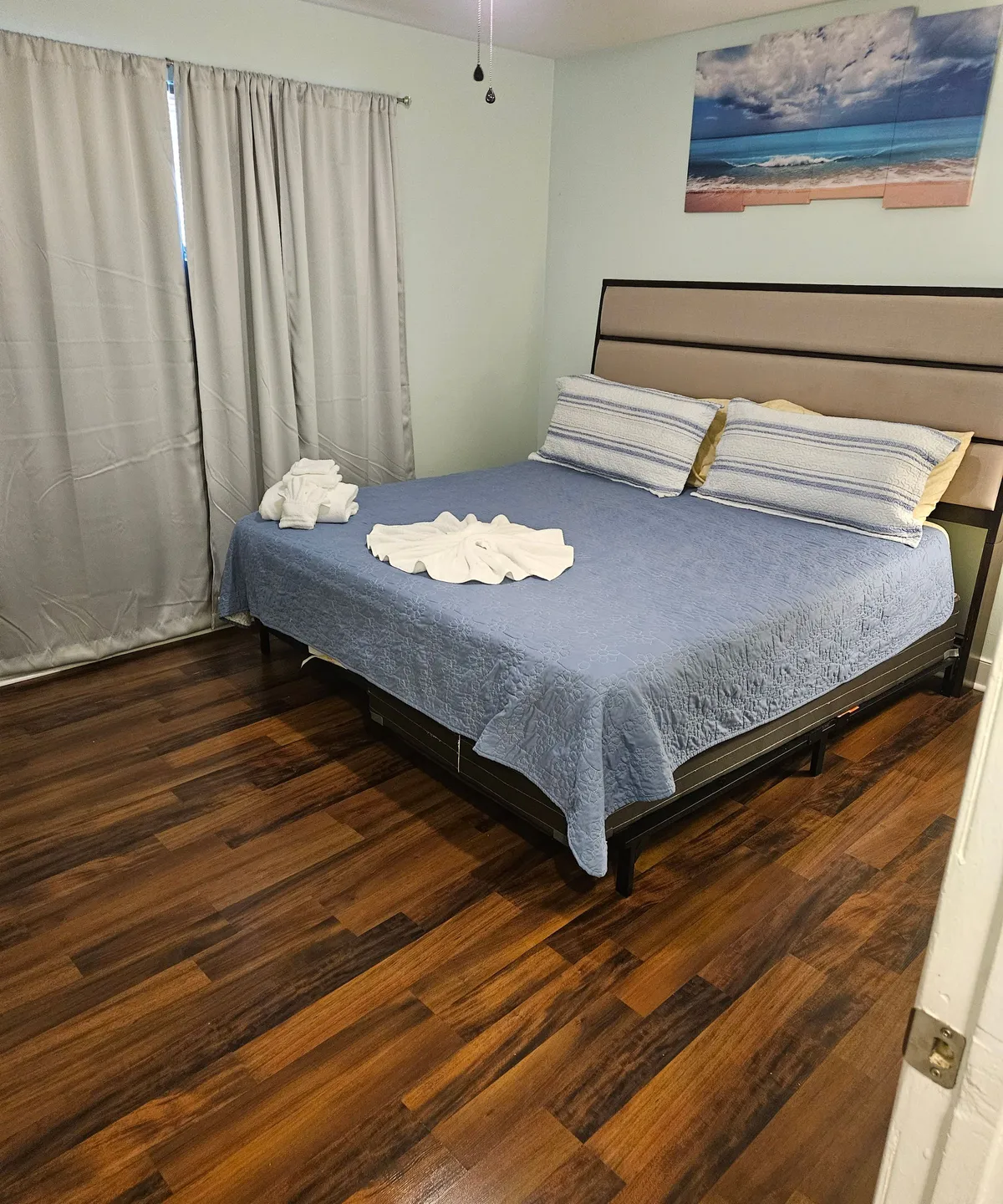 This image features a serene bedroom with a bed adorned in a blue coverlet and decorative pillows, complemented by wall art depicting a calming beach scene and soft curtains letting in natural light. The atmosphere is peaceful and restful, perfect for unwinding after a day out.