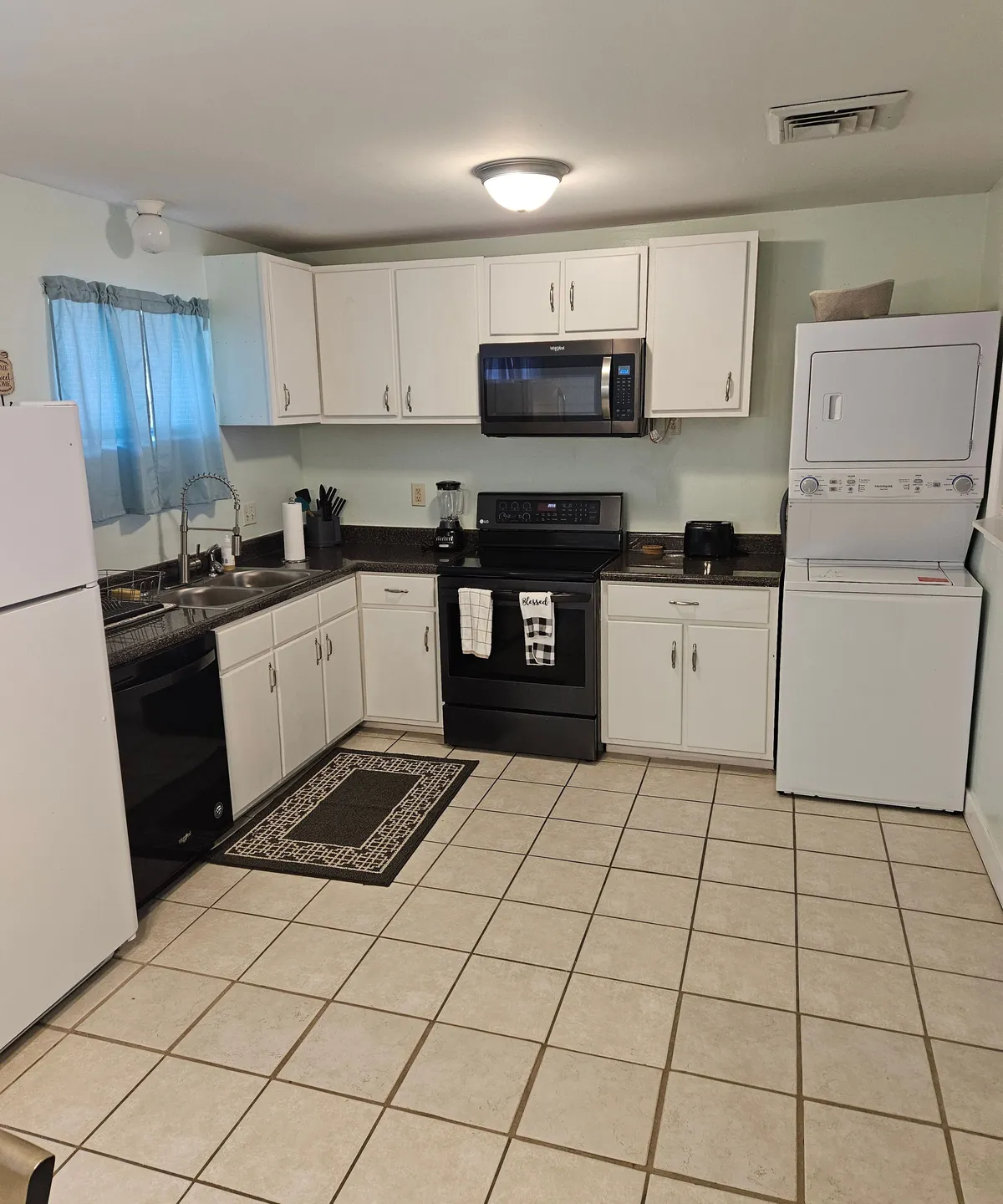 This image showcases a practical kitchenette with black modern appliances, crisp white cabinetry, and a granite countertop. The window with blue curtains brings in natural light, making the space feel inviting. A floor mat adds a cozy touch, while the arrangement of appliances and counter space emphasizes functionality.