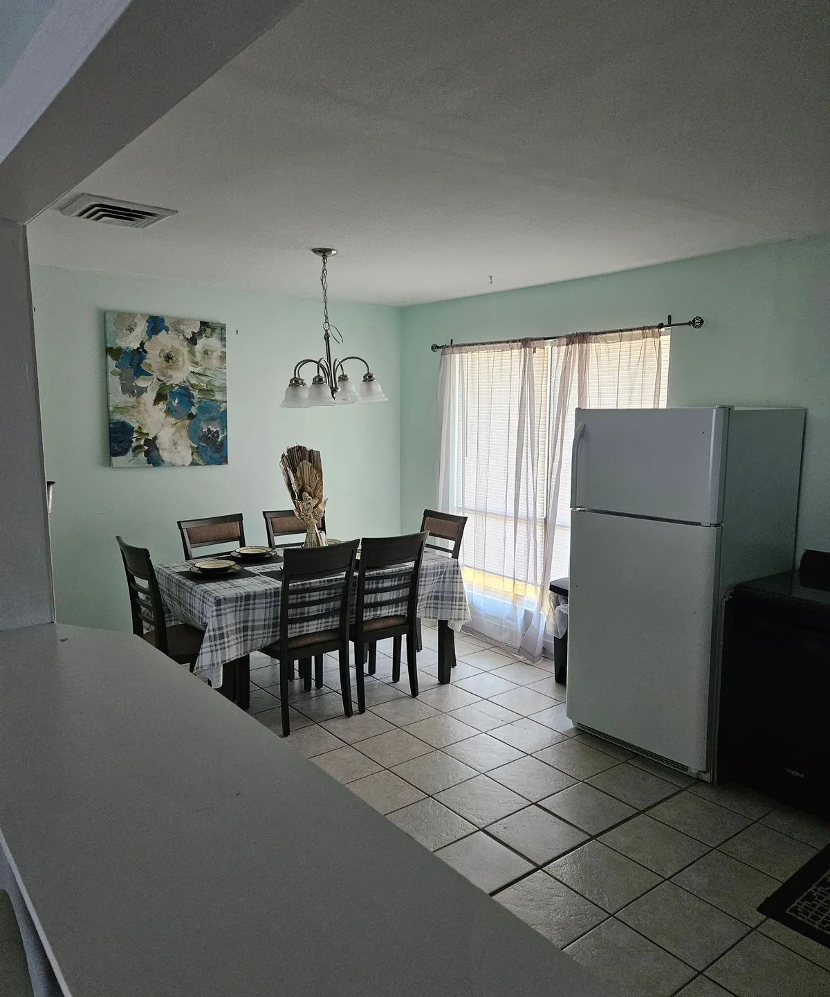 A bright and open dining area with a dining table, wall artwork, refrigerator, and natural light filtering through curtains. The scene is set for family meals or social gatherings, illustrating the heart of the home just off the entryway.