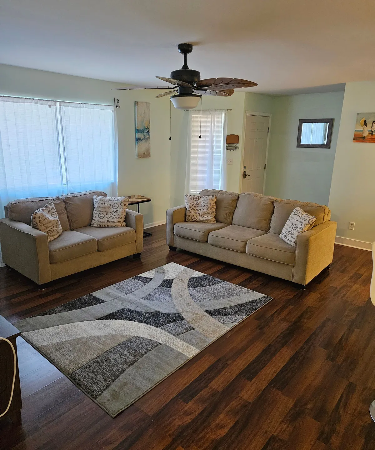 A comfortable living space featuring a plush sofa, stylish rug, ceiling fan, and large windows that fill the room with daylight. The arrangement invites relaxed conversations or quiet reading moments, reinforcing the social, welcoming vibe.